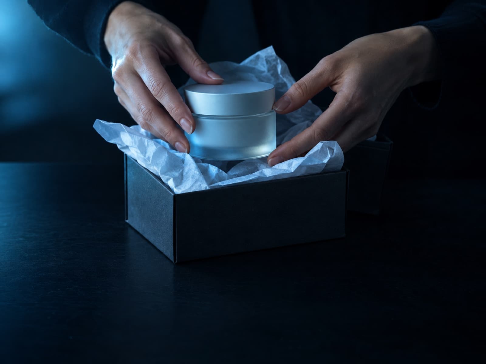 Hands lifting a frosted glass jar out of a gift box lined with tissue paper.
